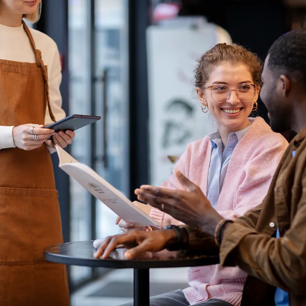 A smiling male waiter chatting to customers at a restaurant table