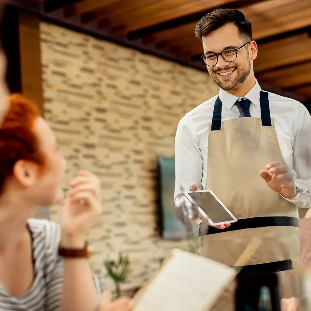 A smiling male waiter chatting to customers at a restaurant table